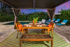 Un espace extérieur aménagé pour les repas, avec une piscine, entouré de verdure et couvert d’une pergola.