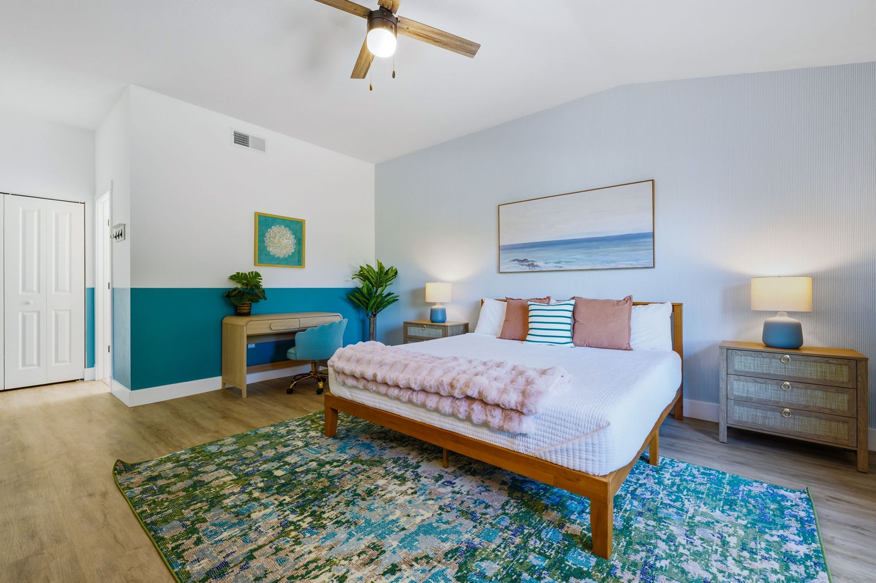 A bedroom with a bed, a desk, a chair, a ceiling fan, a nightstand, a lamp, a painting, and a door.