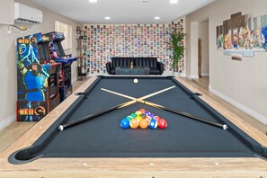 Ein Billardtisch mit Kugeln und Spielstöcken, ein Spielautomat, ein schwarzes Sofa und eine Wand voller Fotos.