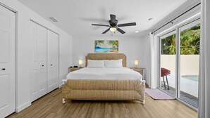 A bedroom with a large bed, a ceiling fan, a painting of a beach, and a view of a pool through a sliding glass door. - Sleeps 6 Private Pool Fire Pit Game House (Sarasota)