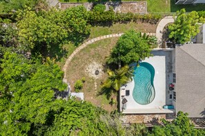 Eine Vogelperspektive zeigt ein Anwesen mit Schwimmbecken, umgeben von üppiger Vegetation und einer gepflasterten Terrassenfläche.