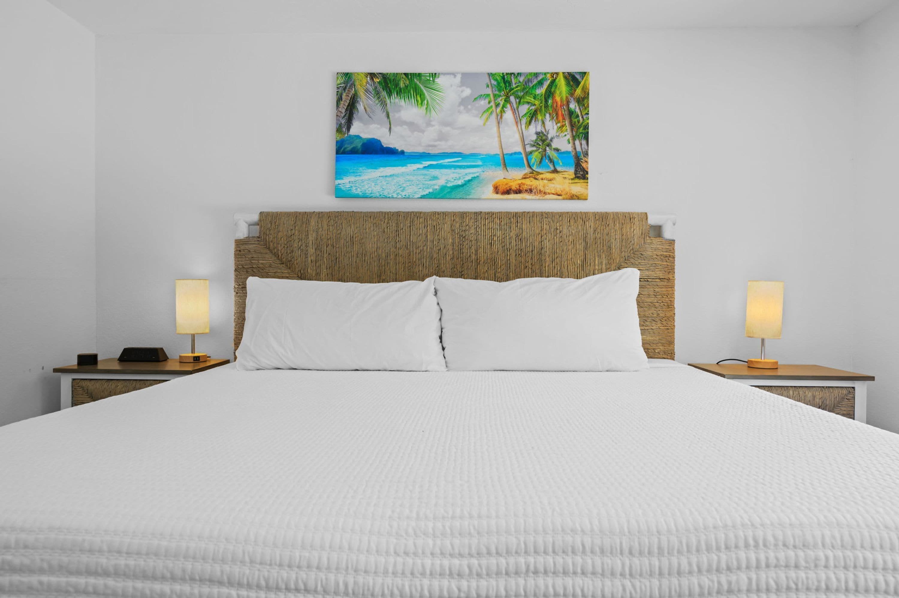 A hotel room with a bed, two bedside lamps, a painting of a beach scene, and a textured headboard.