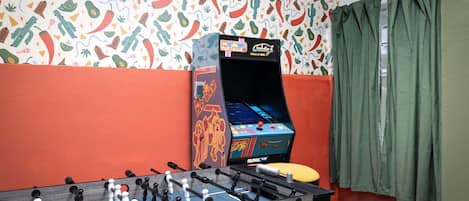 Una habitación con mesa de fútbol, máquina de videojuegos antigua y un patrón de pared colorido.