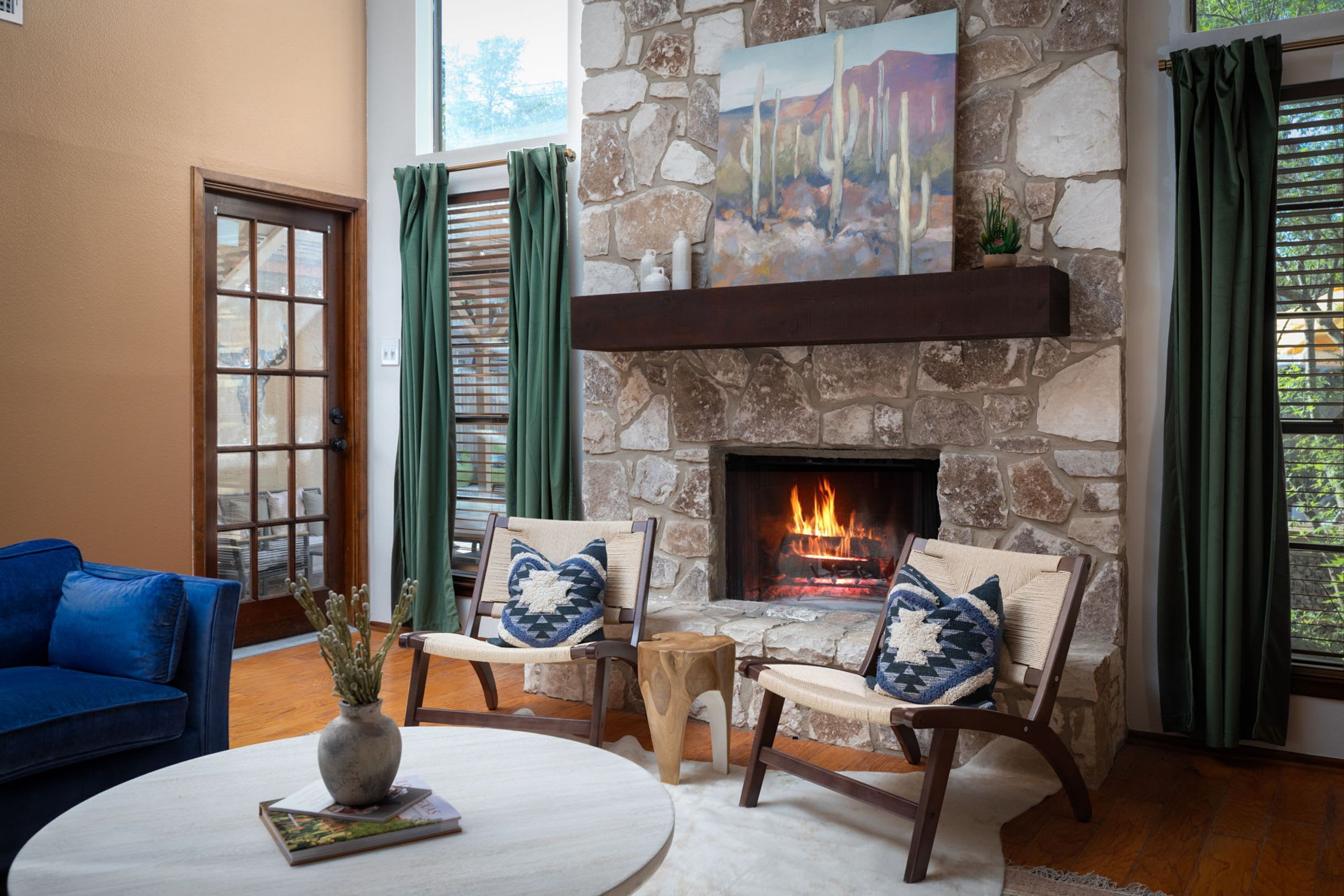 A cozy living room with a stone fireplace, two armchairs with cushions, a blue sofa, and a round coffee table with a vase and books.
