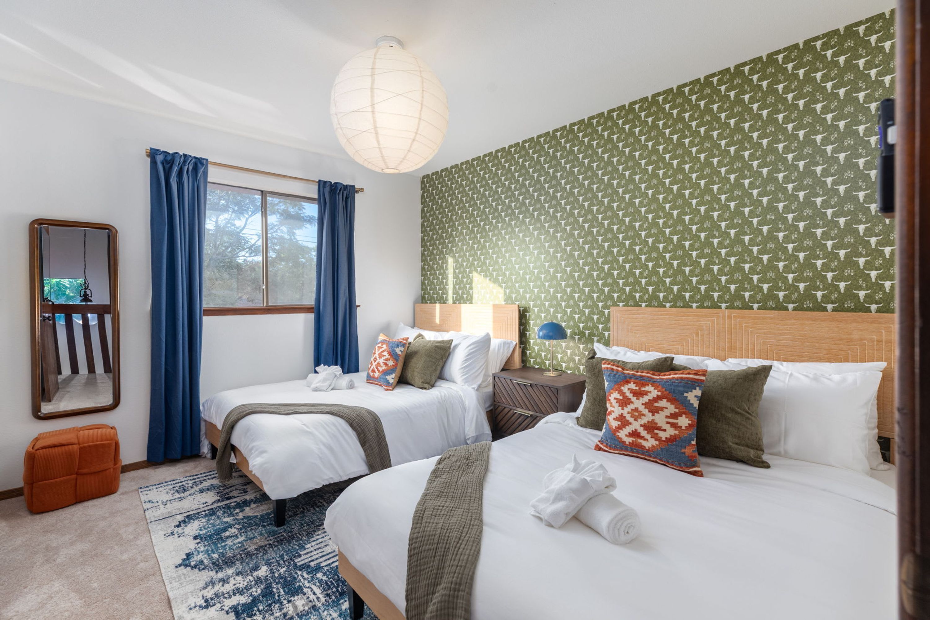 Habitación de hotel con dos camas, un espejo grande, una ventana con cortinas, una pared con diseño y una mesita de noche con lámpara.