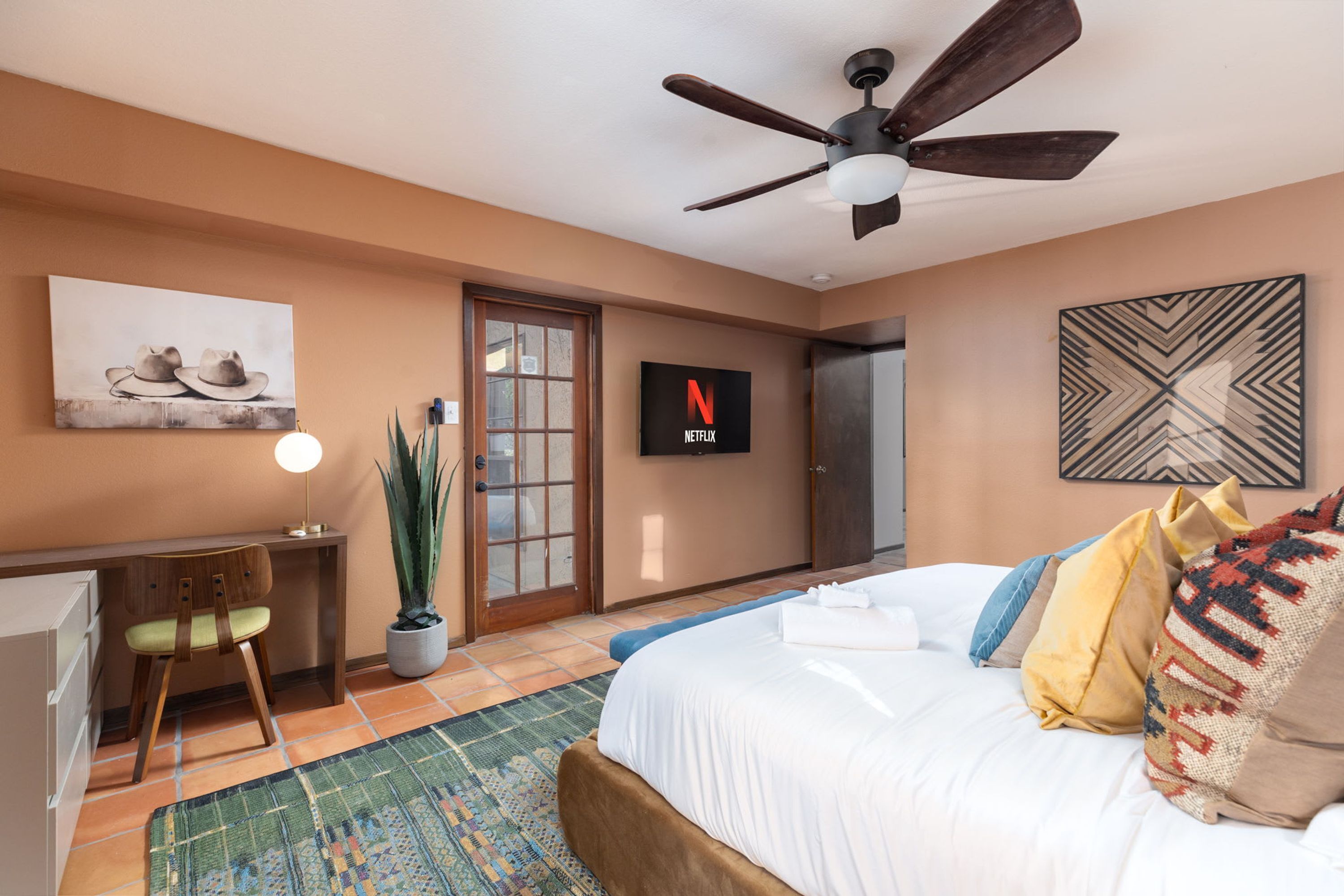 Une chambre à coucher comprenant un lit, un bureau, une chaise, une plante en pot, un ventilateur de plafond et un téléviseur affichant le logo de Netflix.