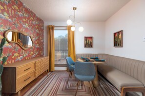 Ein gemütliches Zimmer mit einem hölzernen Schminktisch, einem blauen Sessel, einem runden Tisch mit Teller und Servette, einem Sofa und einem Fenster mit gelben Vorhängen.