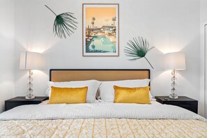 Une chambre d’hôtel avec un lit, deux tables de chevet, deux lampes, un tableau encadré au mur et des feuilles de palmier décoratives.