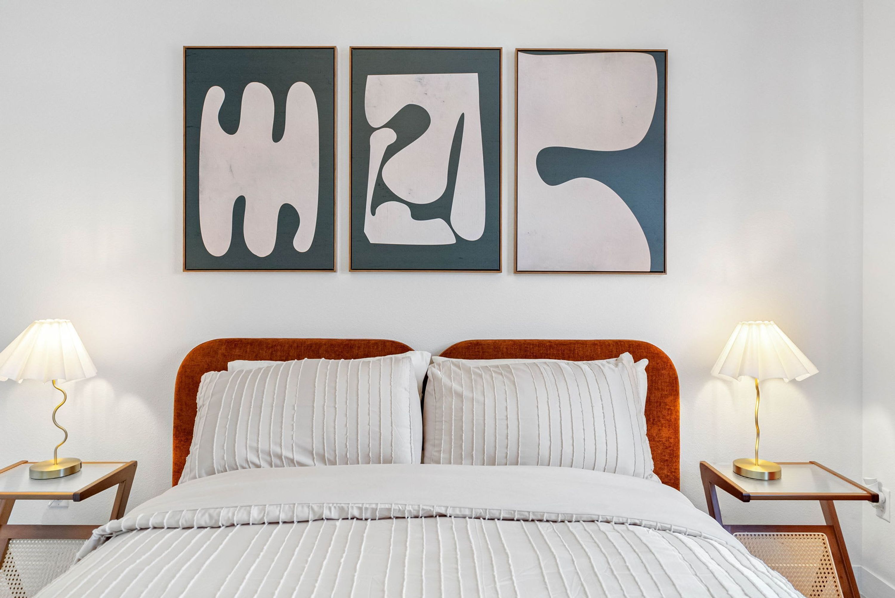 A bed with white bedding and two nightstands with lamps, a headboard, and three abstract wall art pieces.