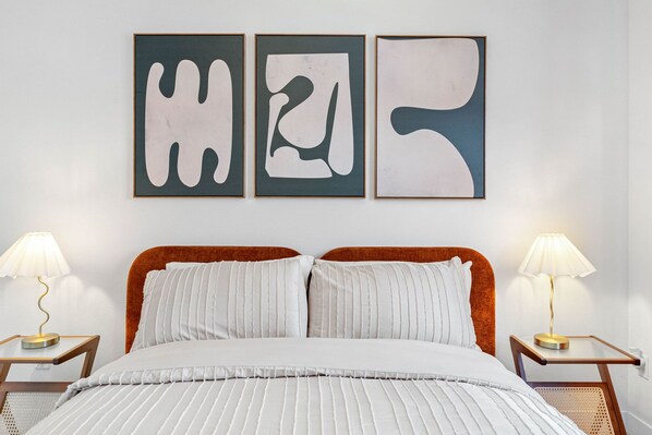 Cama com roupa de cama branca e duas mesas de cabeceira com luminárias, uma cabeceira e três obras de arte abstratas na parede.