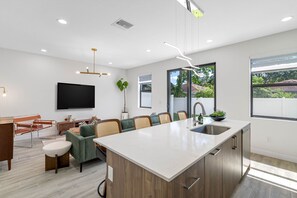 Cozinha moderna com ilha central, área de jantar com sofá e cadeiras, televisão de tela plana na parede e grandes janelas que oferecem luz natural.