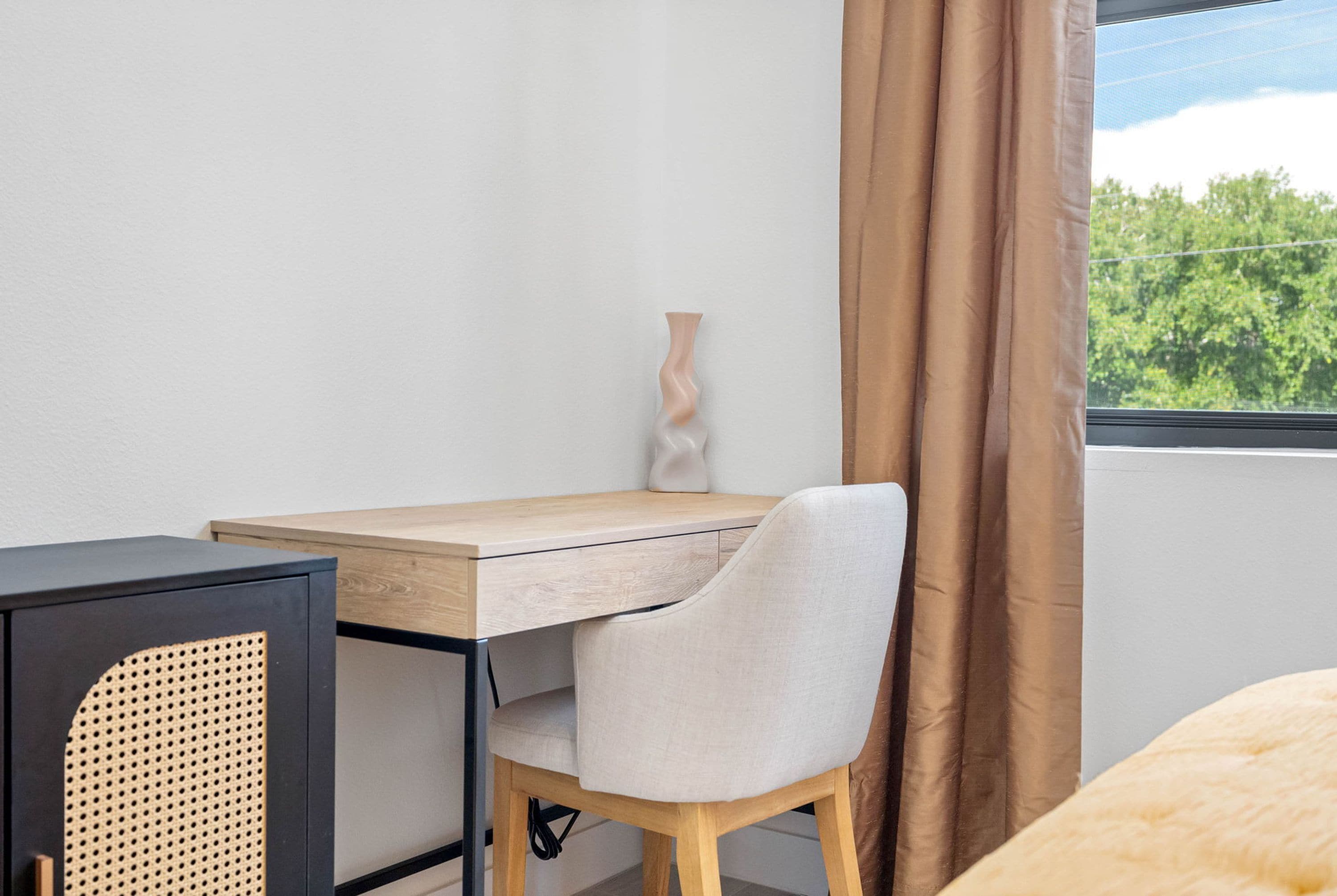 Ein Zimmer mit Schreibtisch, Stuhl und Blick auf Bäume durch ein Fenster.