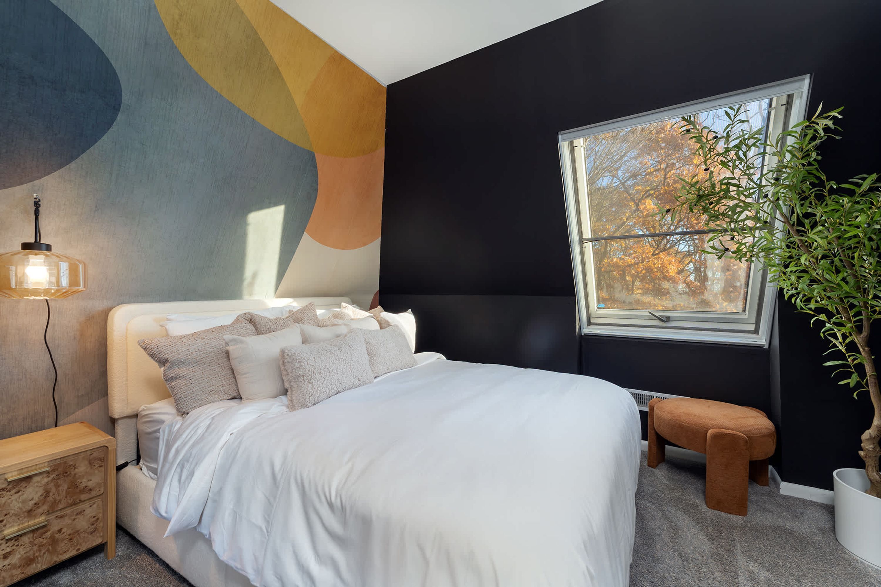 A bedroom with a bed, a bedside table, a lamp, a window with a view of trees, and a plant.