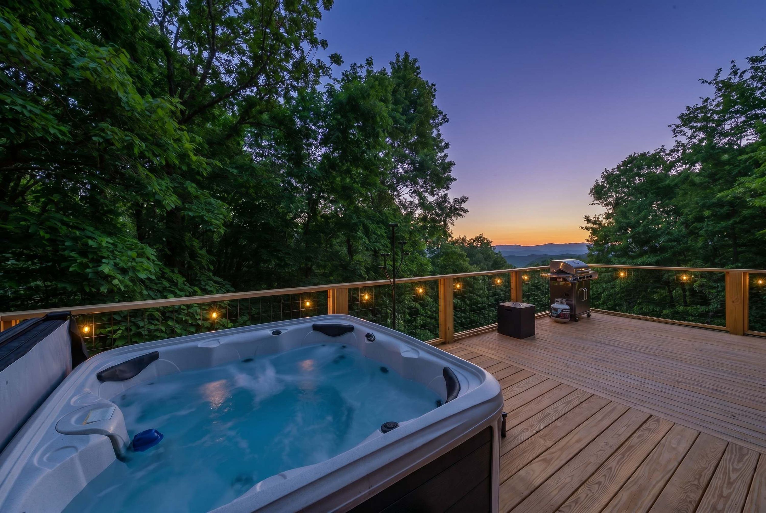A hot tub on a wooden deck with a view of trees and a sunset.