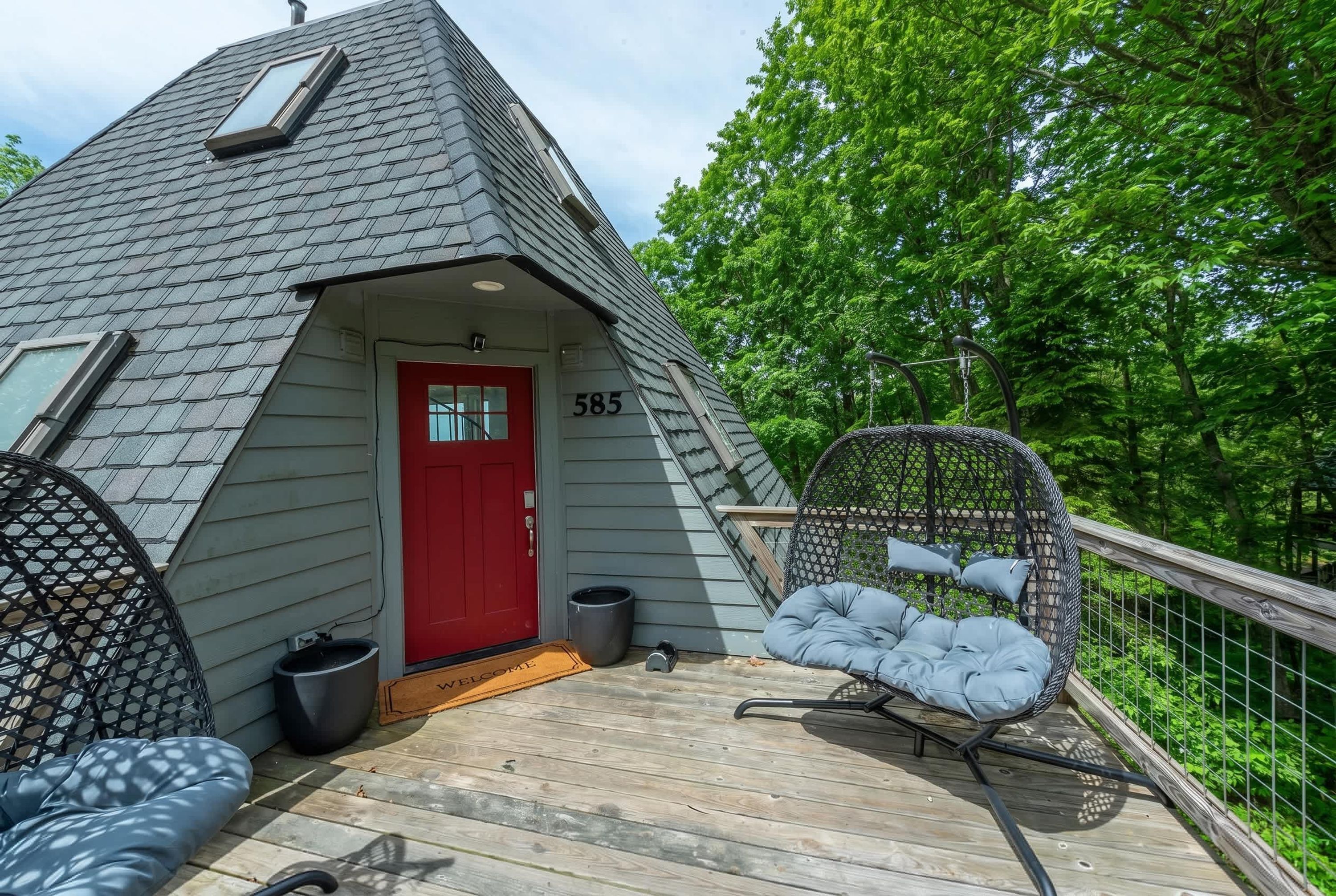 A small, grey, A-frame-style cabin with a red door, numbered 585, featuring a wooden deck, a hanging chair, and a welcome mat.