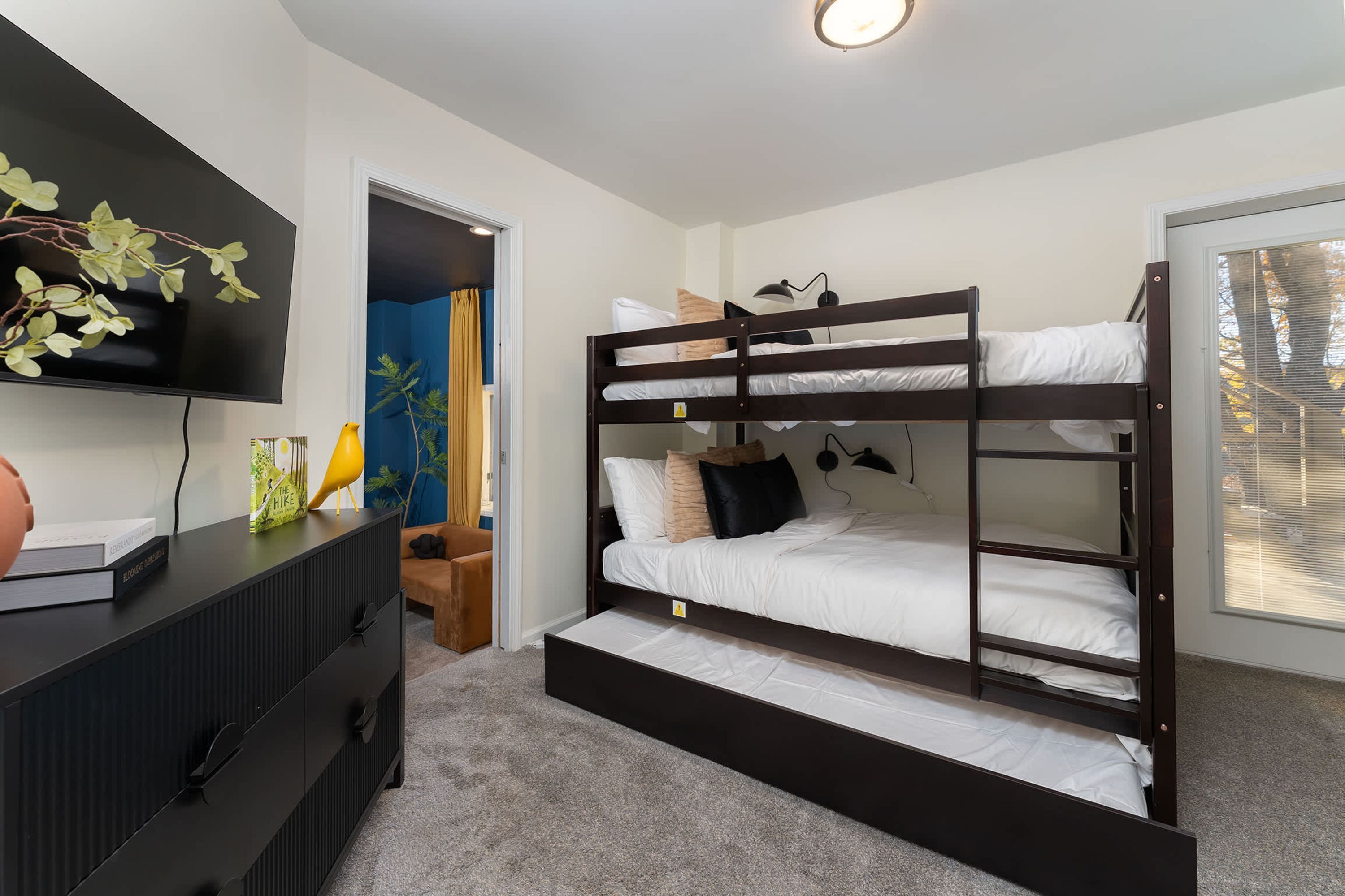 A bunk bed room with a television on the wall, a desk with a lamp, and a window with blinds.