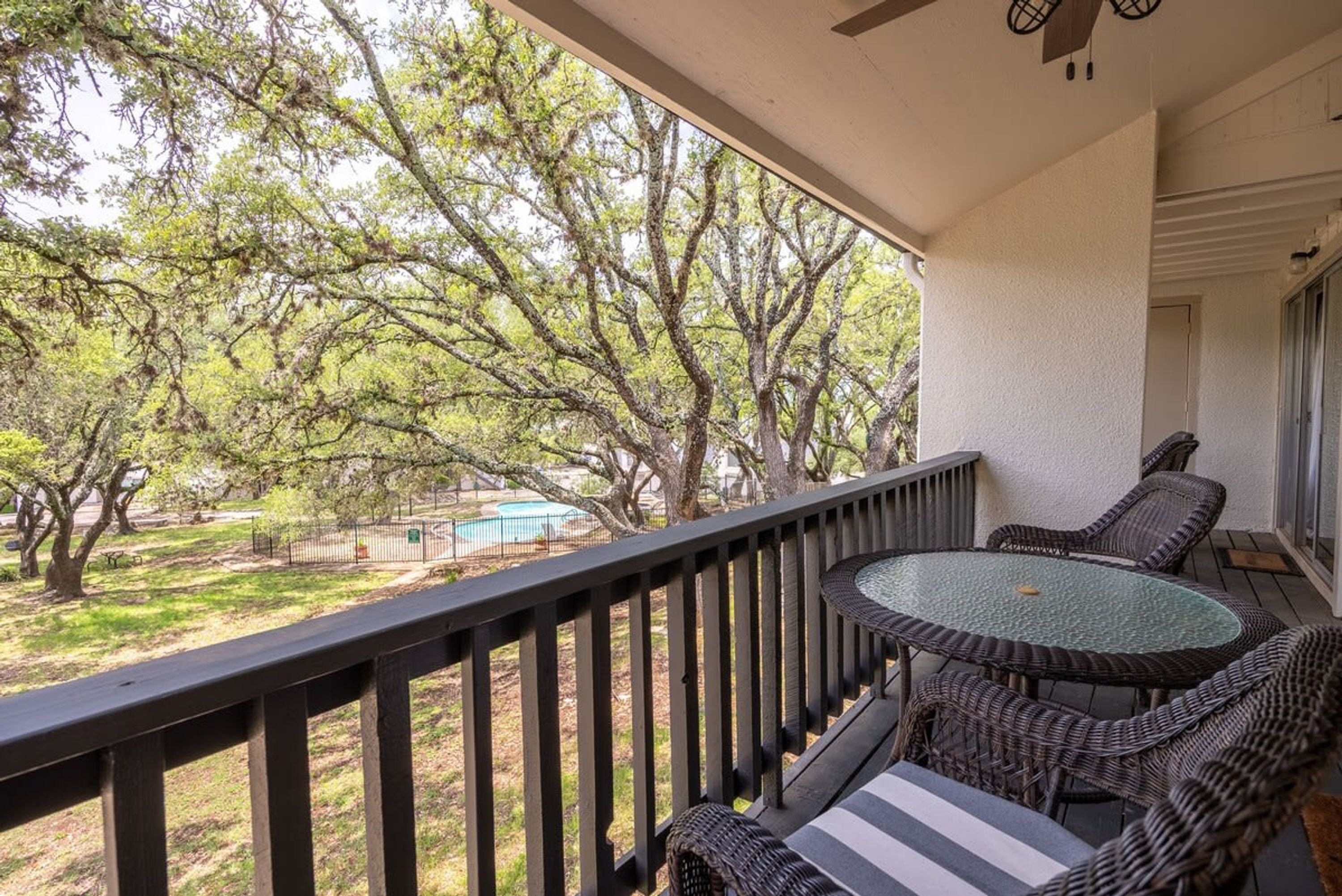 A balcony with wicker furniture, a view of a swimming pool, and trees.