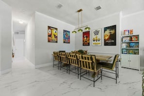 Un comedor con suelo de mármol, una mesa de madera y sillas. Hay carteles en la pared y una estantería con libros y juegos.