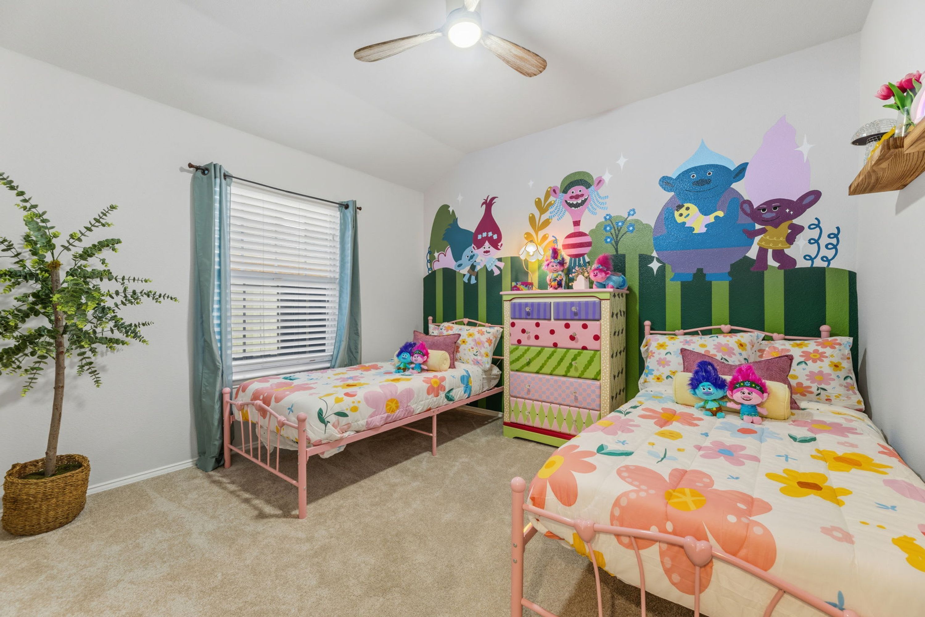 Quarto infantil com duas camas, um penteou e um ventilador de teto, além de uma janela com persianas.