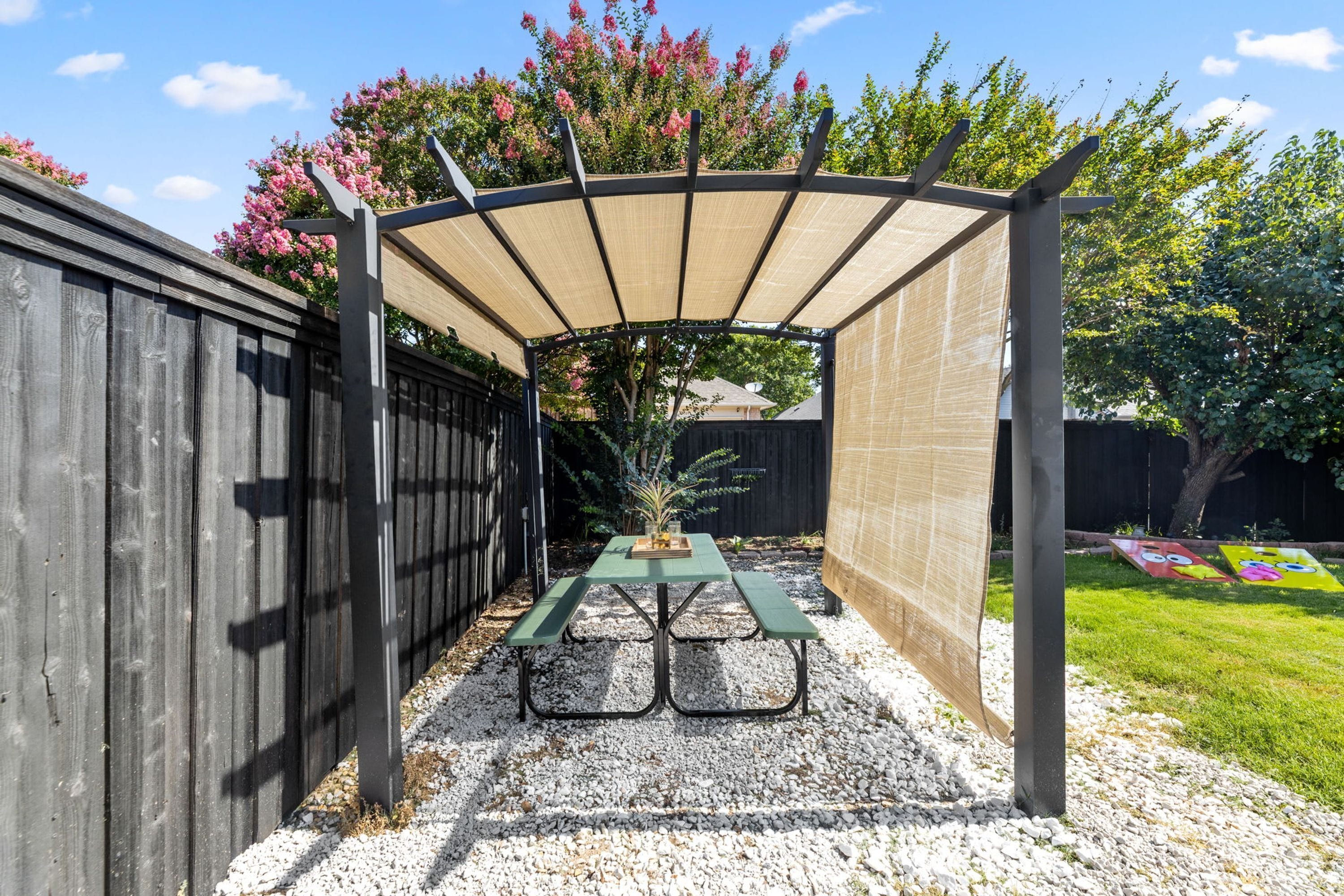 A covered patio area with a table and benches, surrounded by a wooden fence and a garden.