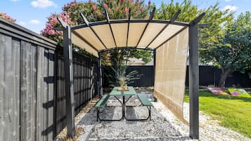 A covered patio area with a table and benches, surrounded by a wooden fence and a garden.