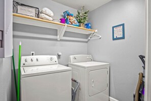 A laundry room with two white washing machines, a green mop, and a shelf with various items including a box labeled 'Body/Paws'. - Copano Ridge Private Pool Priv Dock Pet Friendly (Rockport)