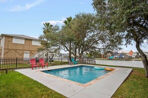 Uma piscina retangular com borda azulejada, rodeada por um deck de concreto. Há duas cadeiras Adirondack vermelhas e um bote inflável azul.