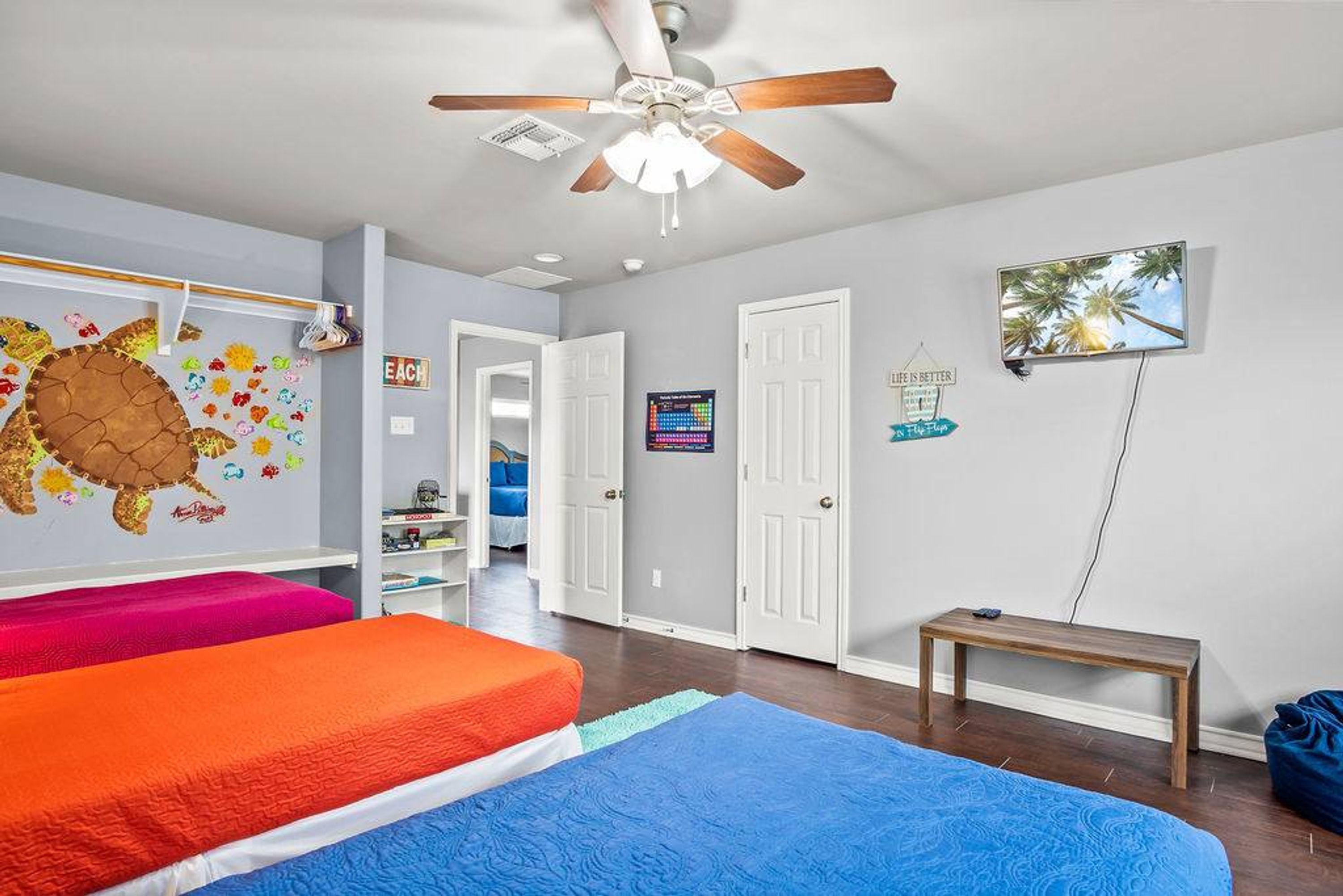 A room with two beds, a ceiling fan, a TV, and a door leading to another room.