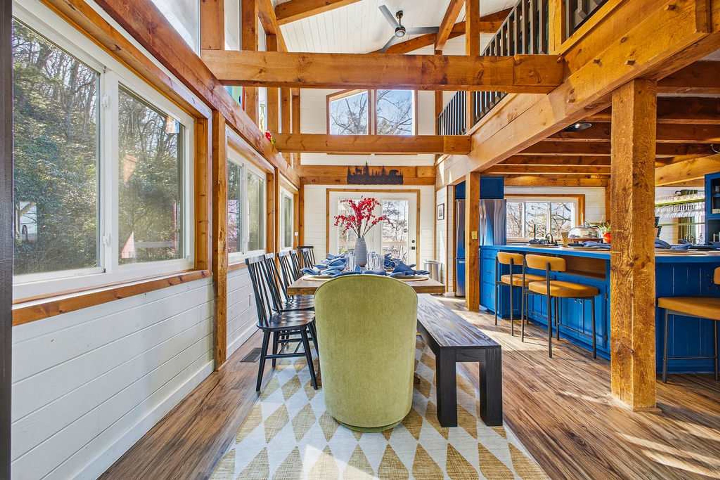 A spacious dining area with a high ceiling and wooden beams, a bar area with blue bar stools, and a view of the outdoors through large windows.