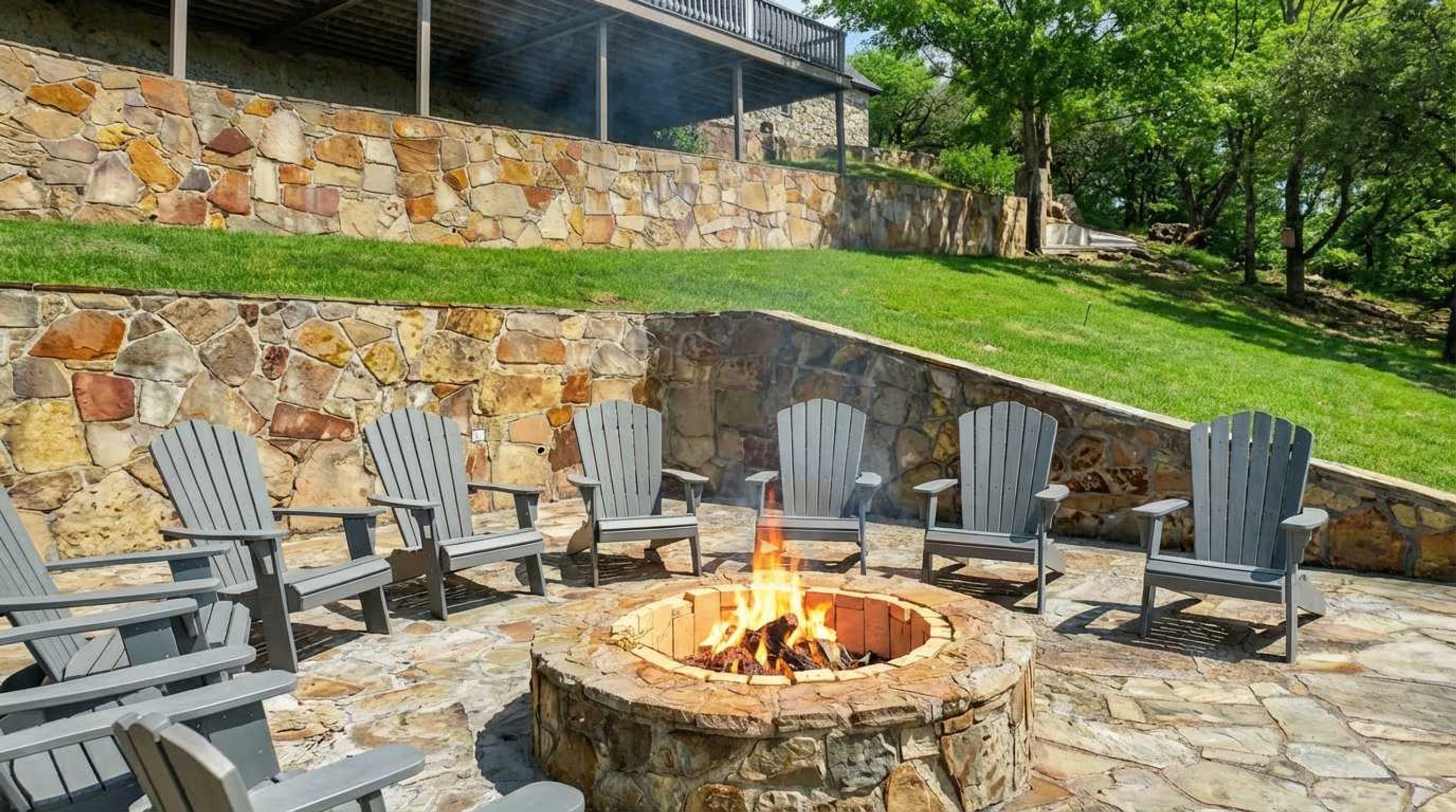A stone fire pit with chairs around it, set against a stone wall with a grassy slope behind.
