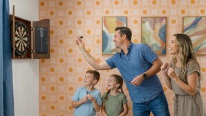 Una familia jugando dardos en una habitación con papel tapiz estampado y arte abstracto en las paredes.