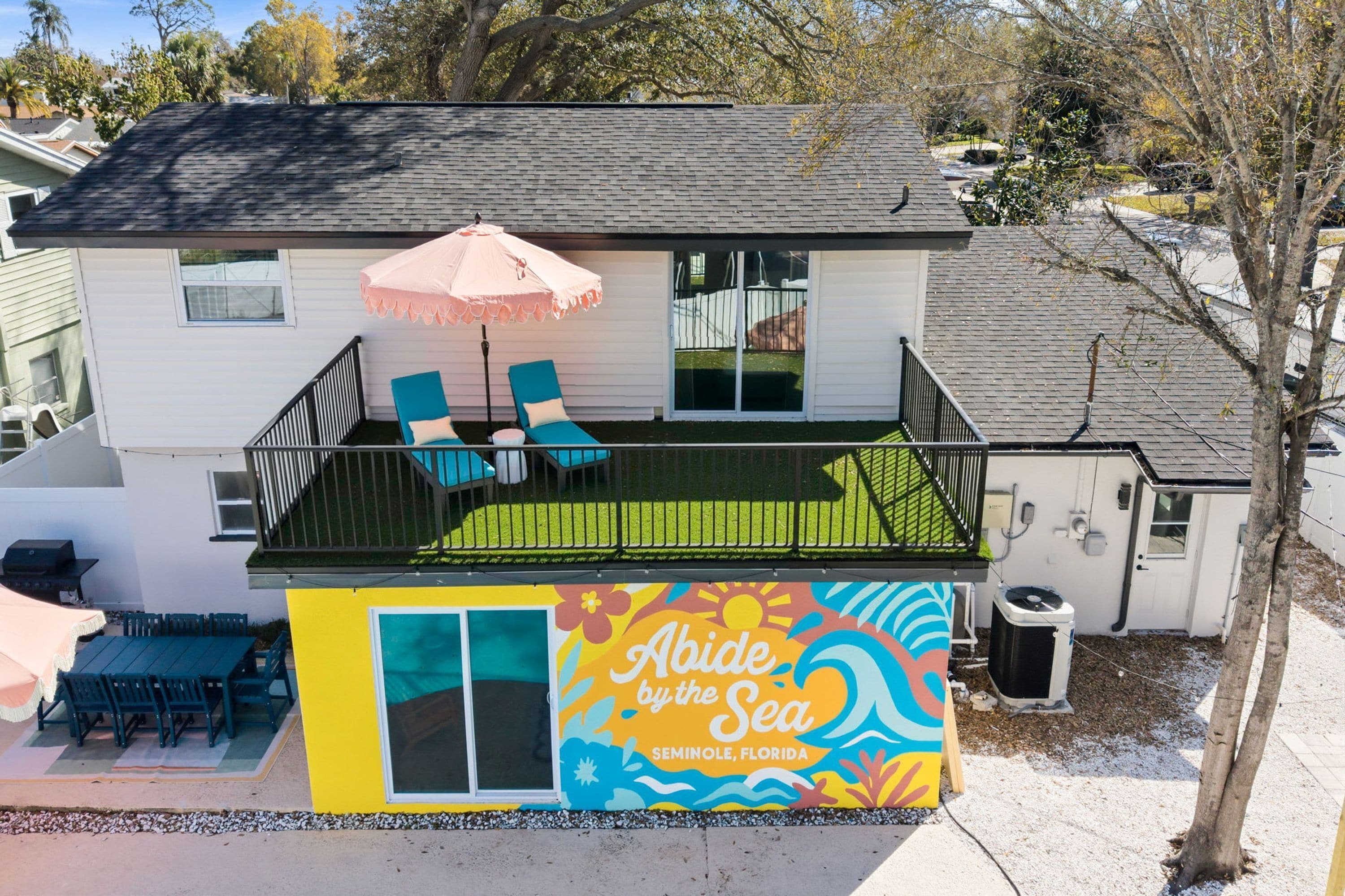 A two-story house with a balcony, a patio with chairs and an umbrella, and a colorful mural on the side.