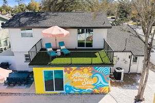 Una casa de dos pisos con un balcón, un patio con sillas y un parasol, y un mural colorido en la pared.