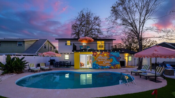 Un área de piscina con sillones reclinables, un edificio pequeño con un mural y una bandera.
