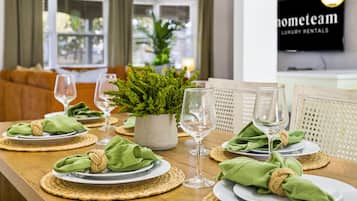 A dining table set with plates, napkins, and wine glasses, with a potted plant and a television in the background.