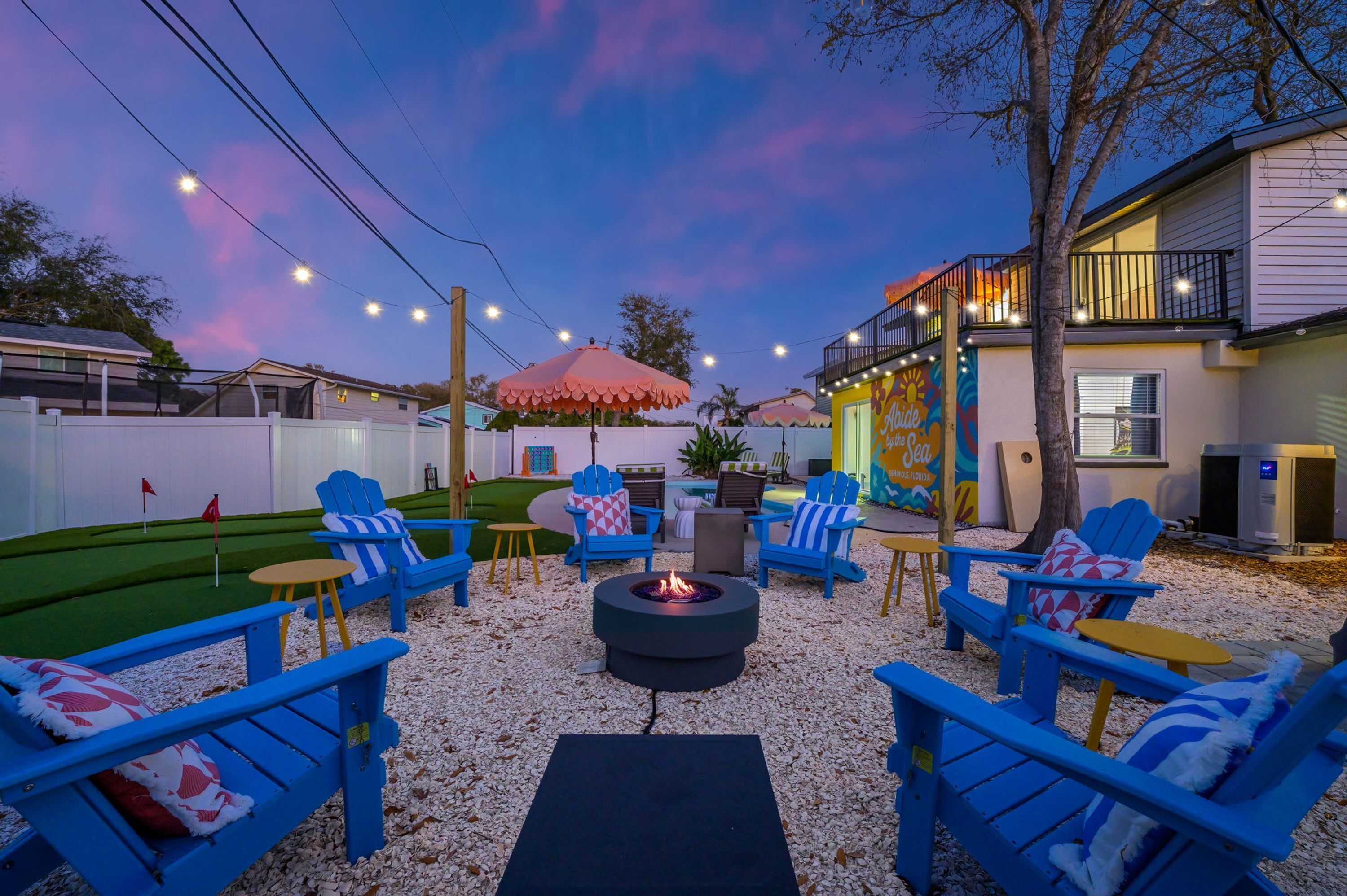 Un espacio al aire libre con sillas azules y una chimenea, rodeado por una cerca blanca y luces colgantes.