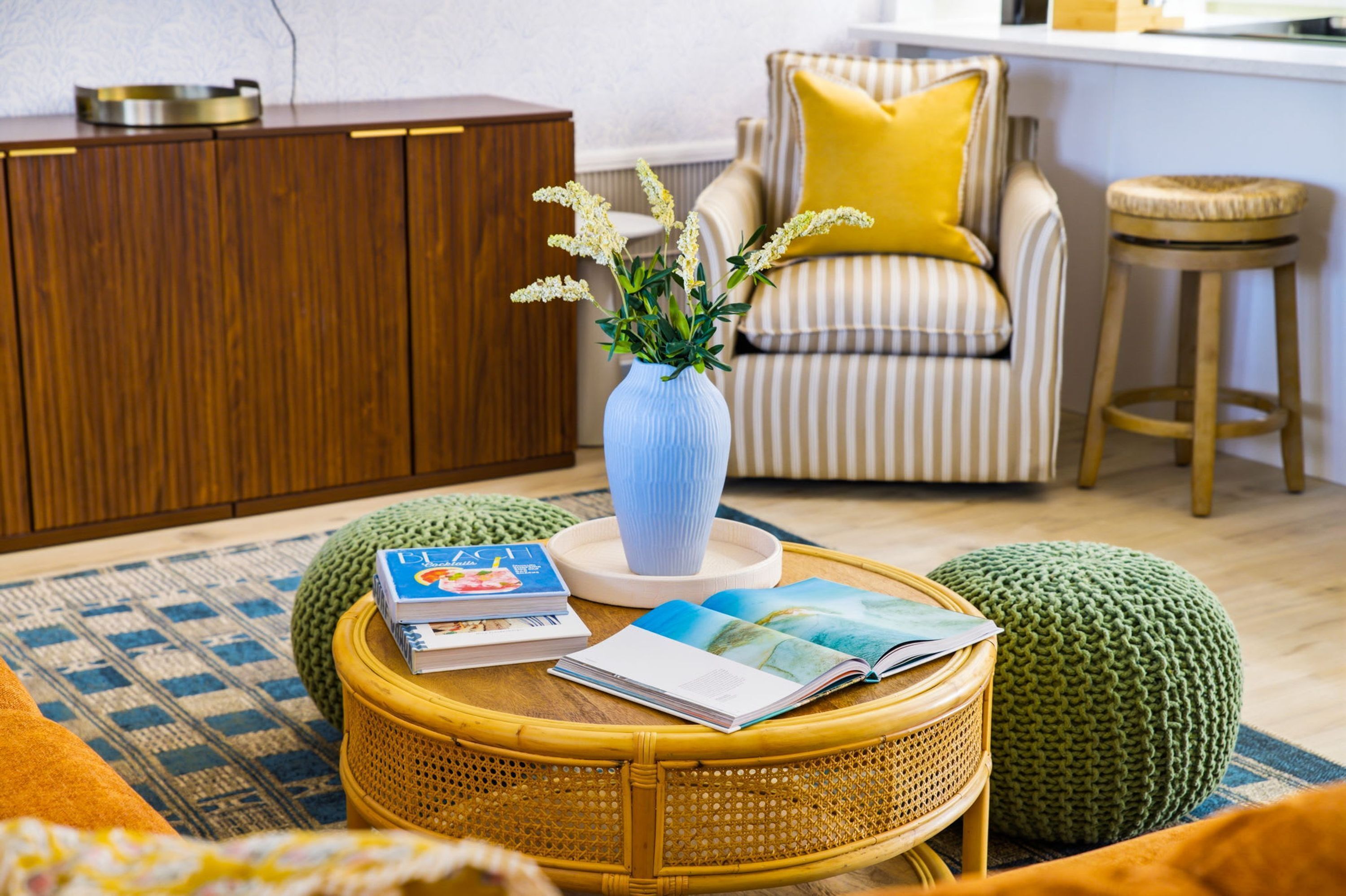 Una sala de estar con una mesa de centro de mimbre, un sillón a rayas con un cojín amarillo, un aparador de madera y un jarrón con flores.