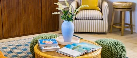Una sala de estar con una mesa de centro de mimbre, un sillón a rayas con un cojín amarillo, un aparador de madera y un jarrón con flores.