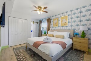 Un dormitorio con una cama, un ventilador de techo, una mesita de noche, una lámpara y un muro con obras de arte enmarcadas.