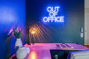 Ein Zimmer mit einer Neonreklame, die 'OUT OF OFFICE' zeigt, einem Tisch mit Lampe und einer Pflanze.