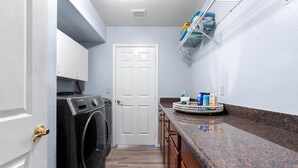 A laundry room with a front-loading washer and dryer, a white door, and a countertop with cleaning supplies. - Epic Family Getaway Sport Court Heated Pool (Scottsdale)