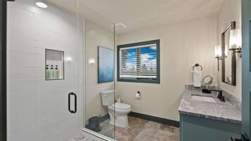 A bathroom with a glass shower enclosure, a toilet, a sink with a marble countertop, and a window with blinds.