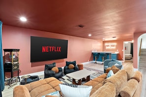 A living room with a large flat-screen TV mounted on the wall displaying 'NETFLIX' in red letters, a sofa set, a coffee table, and a kitchen area in the background. - AZ Game House Sport Court - Golf - Pool - Games (Peoria)