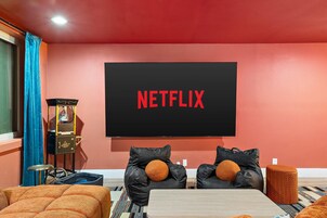 Una stanza con una grande TV a schermo piatto montata su una parete rossa, due divani in pelle nera con cuscini arancioni, un tavolino in legno e una macchina per popcorn d'epoca.