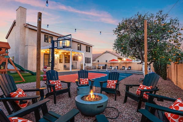 An outdoor seating area with Adirondack chairs and a fire pit, a basketball court, and a house with a visible basketball hoop. - AZ Game House Sport Court - Golf - Pool - Games (Peoria)
