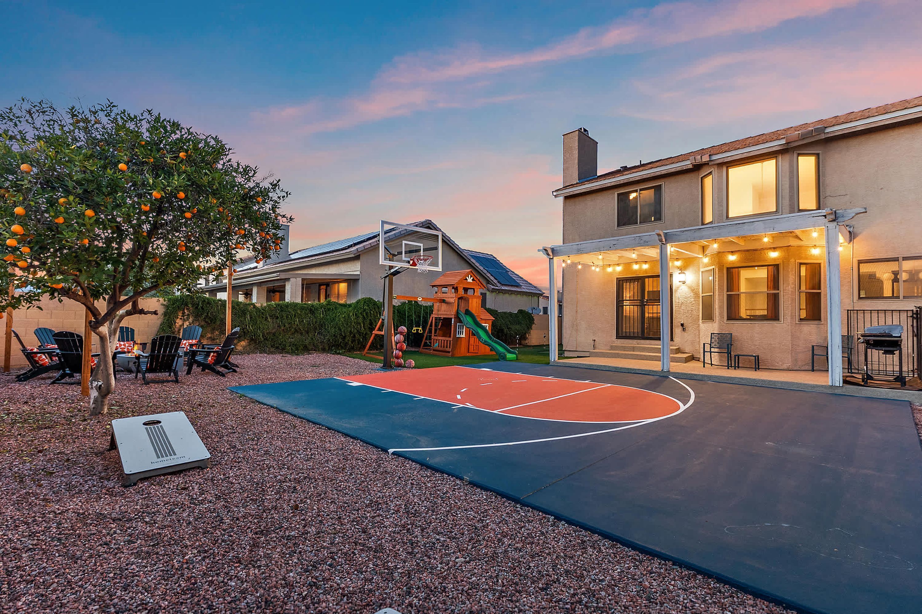A basketball court with a slide, surrounded by a gravel area and a tree with fruit.