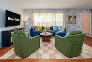 Ein Wohnzimmer mit einem blauen Sofa, grünen Sesseln, einem Couchtisch und einem Fernseher an der Wand.