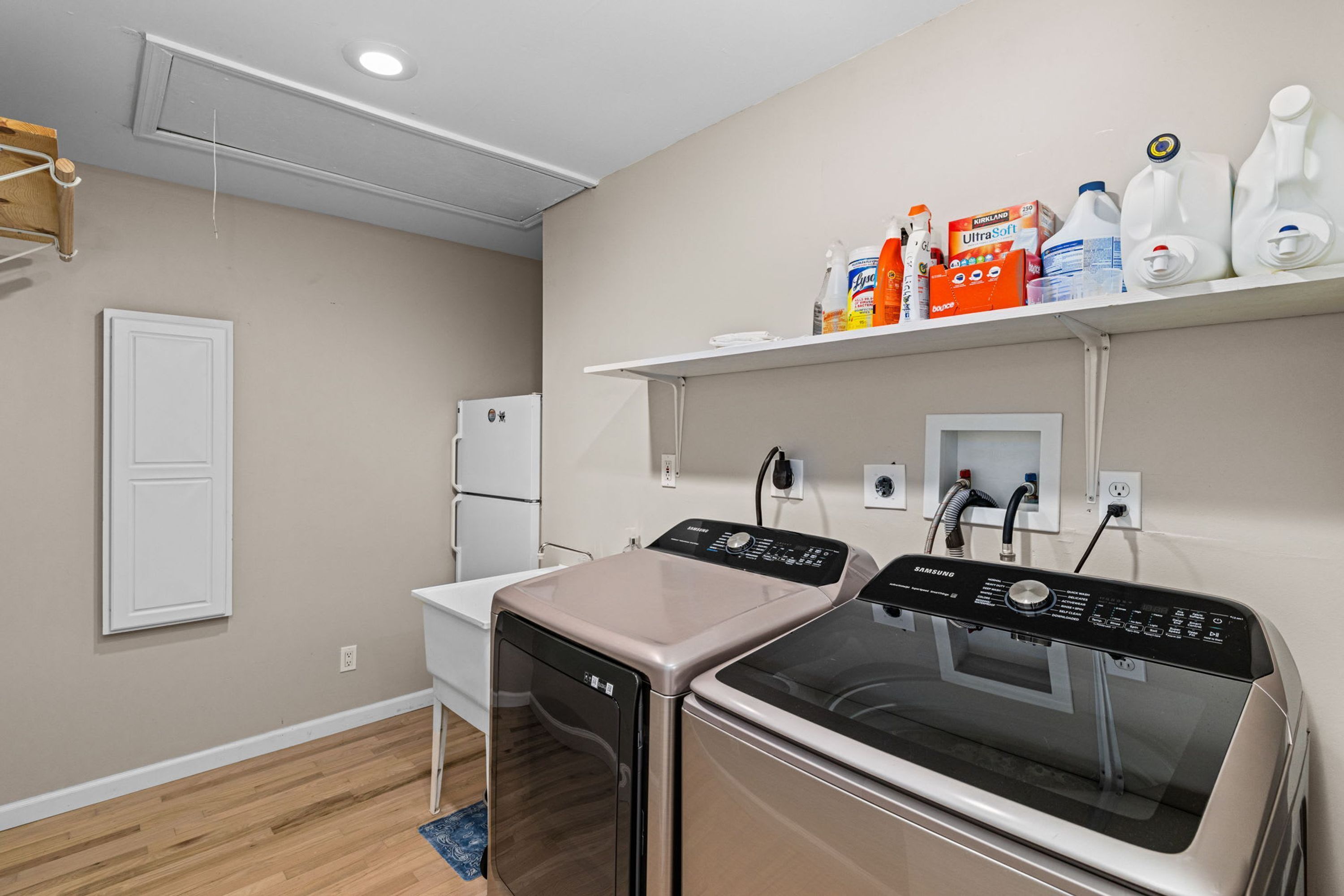 A laundry room with two washers and dryers, a white refrigerator, and a shelf with cleaning supplies.