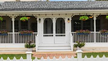 A two-story house with a white picket fence, a front porch with potted plants, and a clear blue sky.