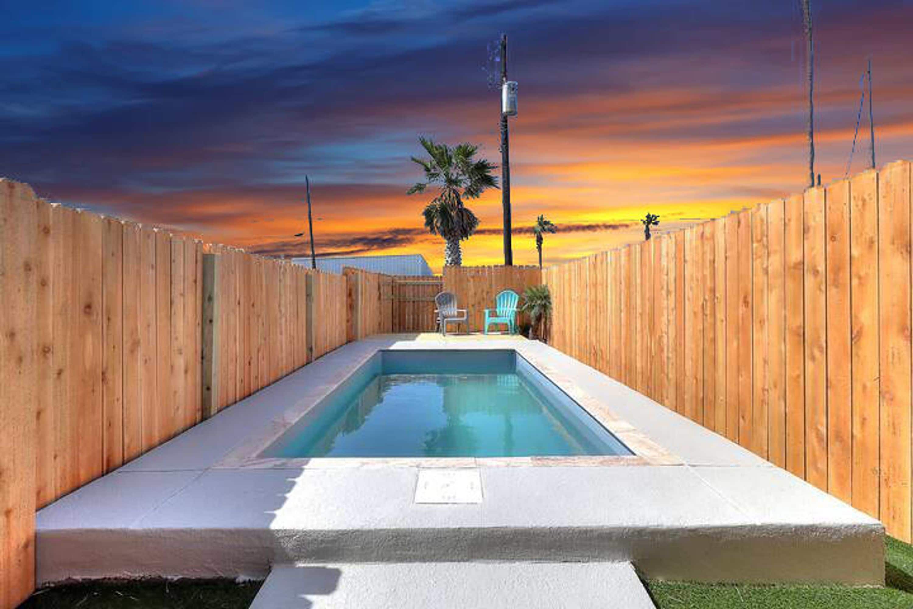 A rectangular swimming pool with a wooden fence surrounding it, a palm tree, and a clear sky.